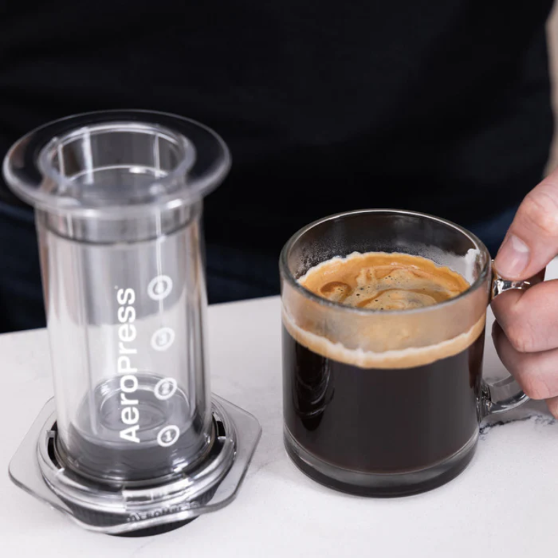 AeroPress Clear plunger stående ved siden af en glasespresso — klar AeroPress-plunger på bordet ved siden af en lille glas med espresso og cremalag; hånd i billedets højre side løfter koppen, stilrent køkkenbaggrund og neutralt bord.