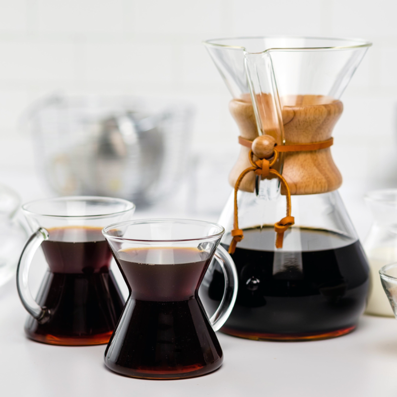 Her ses en Chemex Classic 8 kops med nybrygget kaffe i. Foran står der to glas formet som Chemex kander med hank, i dem er der kaffe.