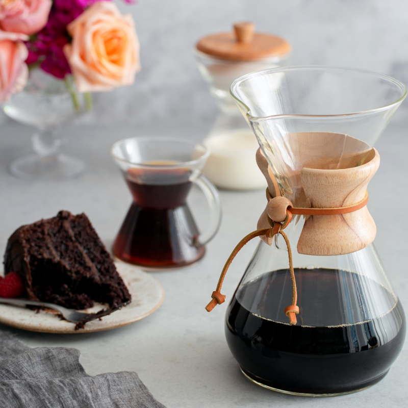 Her ses en Chemex Classic 8 kops med nybrygget kaffe i. Bag ved står der 1 glas formet som Chemex kander med hank, med kaffe i. Ved siden af glasset er en tallerken med et stykke chokoladekage på. Bagerst kan man se Chemex mælkekande med mælk i. I venstre hjørne er der en buket roser.