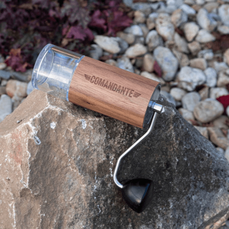 En Comandante C40 kaffekværn med træfinish ligger på en stor sten omgivet af småsten.