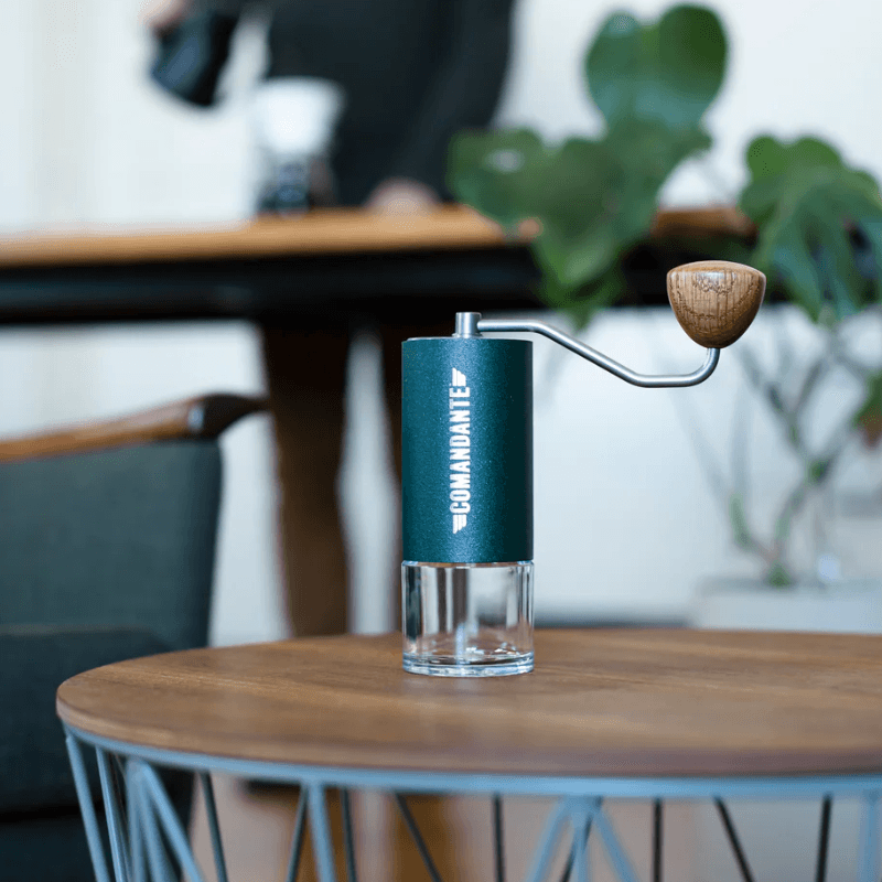 Grøn Comandante C40 kaffekværn står på et lille sofabord med en person og planter i baggrunden.