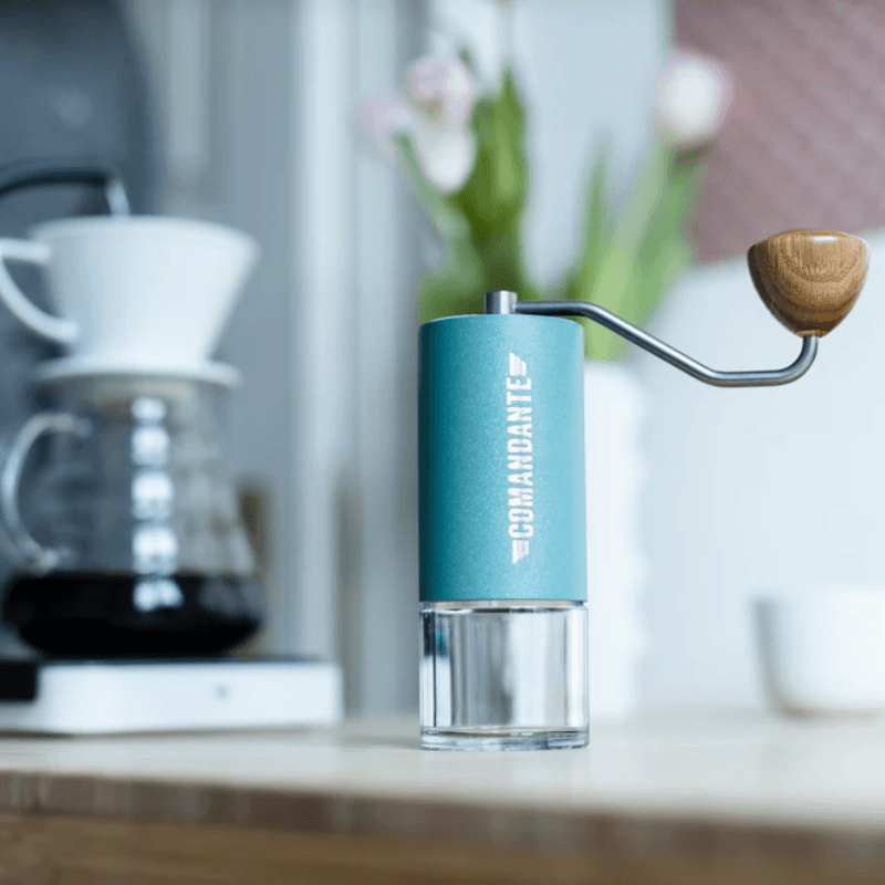 Turkis Comandante C40 kaffekværn placeret på et køkkenbord med en kaffebrygger og blomster i baggrunden.