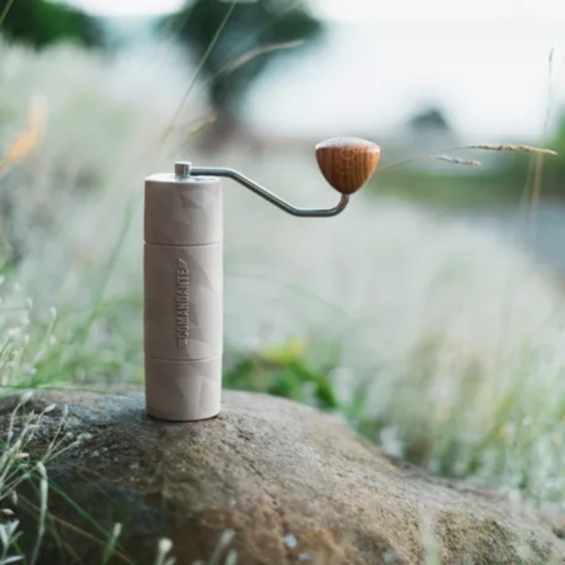 Comandante X25 kaffekværn placeret på en sten i et udendørsmiljø, omringet af højt græs. Kværnen illustreres som et bærbart og praktisk redskab til udendørs kaffeentusiaster.