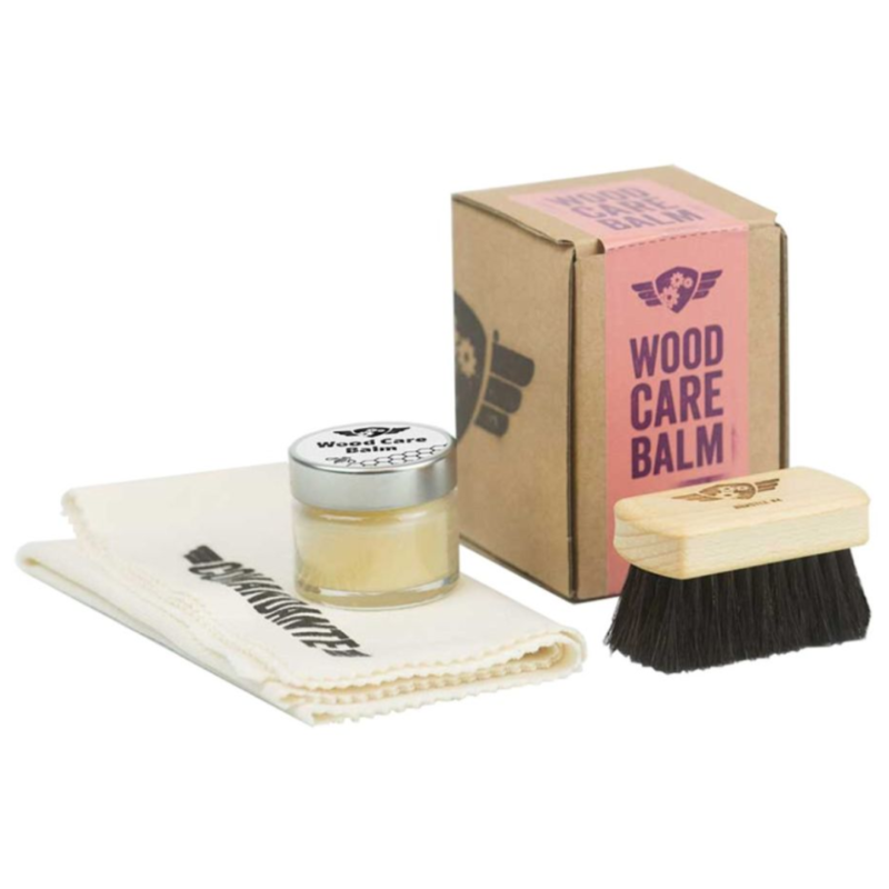 Comandante Wood Care Balm sæt, som består af en lille glaskrukke med et metal låg, der indeholder en speciel træplejebalsam. Krukken har en sort etiket på låget med teksten "Wood Care Balm" og Comandante-logoet. Sættet inkluderer også en blød, foldet rengøringsklud med "Comandante"-logoet trykt på. Ved siden af ses en træbørste med sorte børstehår og et indgraveret Comandante-logo på træhåndtaget. Det hele præsenteres med en brun papæske, der bærer en lyserød etiket med teksten "Wood Care Balm" og logoet.