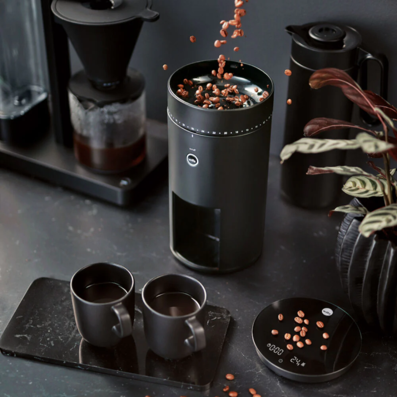 Wilfa Uniform kaffekværn i brug, hvor kaffebønner hældes ned i kværnen, med to kopper kaffe ved siden af.