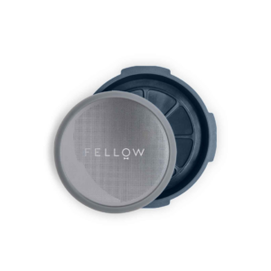 En åben Fellow Prismo AeroPress-adapter, hvor metalfiltret og låsemekanismen tydeligt ses. Adapteren har et minimalistisk design med Fellow-logoet præget på det runde metalfilter.