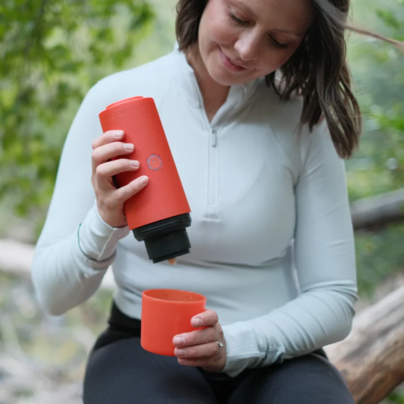 Kvinde nyder friskbrygget espresso i skoven med den bærbare røde PrimeBrew kaffemaskine, ideel til outdoor-eventyr.