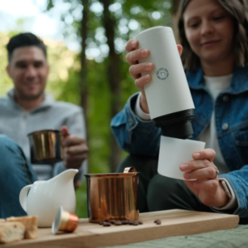 Hvid PrimeBrew kaffemaskine i brug under en hyggelig picnic, mens et par nyder en varm kop espresso i det fri.