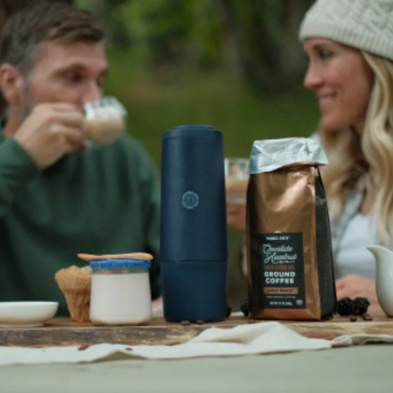 Nomious PrimeBrew i mørkeblå står på et rustikt træbord sammen med kaffebønner, kopper og en pose friskmalet kaffe, skaber en luksuriøs kaffeoplevelse.