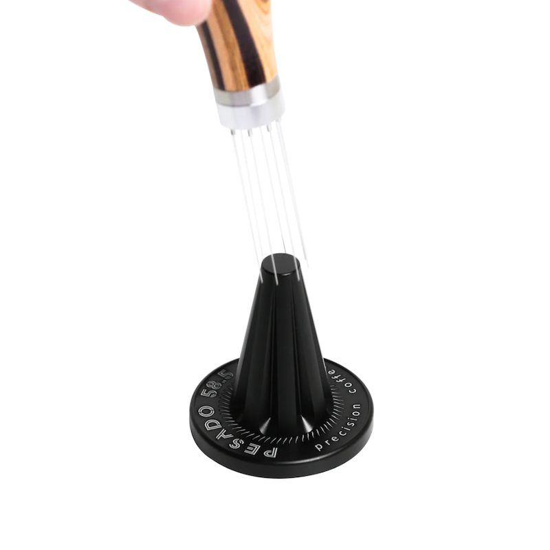 Pesado 58.5 WDT Tool med håndtag i træ og præcisionsnåle, som er ved at blive placeret i standen. Perfekt til ensartet fordeling af kaffegrums.