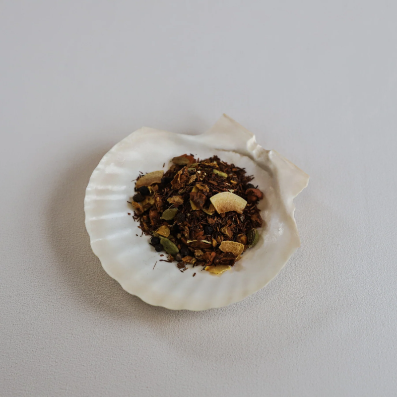 Løse teblade fra Teministeriet No. 730 Rooibos Coconut Ginger te præsenteret i en hvid muslingeskal. Teen består af små, rødbrune rooibosnåle blandet med tørrede kokosflager, grønne kardemommefrø, sorte peberkorn og små stykker ingefær og kanel. Muslingeskallen ligger på en lys overflade, og billedet har en naturlig og æstetisk præsentation, der fremhæver teens unikke ingredienser og teksturer.