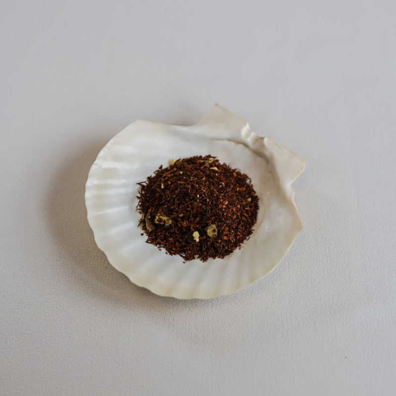 Løse teblade fra Teministeriet No. 731 Rooibos Licorice te præsenteret i en hvid muslingeskal. Teen består af små, rødbrune rooibosnåle blandet med lysebrune lakridsstykker. Muslingeskallen ligger på en lys overflade, og billedet har en ren og naturlig æstetik, der fremhæver teens fine ingredienser og tekstur.