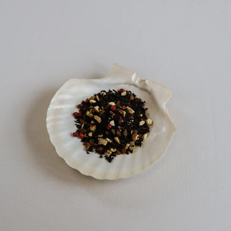 Løse teblade fra Teministeriet No. 531 Black Vanilla Chai te præsenteret i en hvid muslingeskal. Teen består af mørke sorte teblade blandet med små stykker tørret ingefær, kanel, nelliker og røde peberkorn, som giver en krydret chai-karakter. Muslingeskallen ligger på en lys overflade, og billedet har en ren og naturlig æstetik, der fremhæver teens krydrede ingredienser og tekstur.