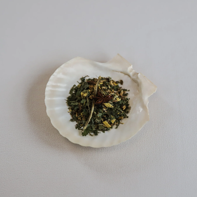 Løse teblade fra Teministeriet Ayurveda Tummy te præsenteret i en hvid muslingeskal. Teen består af tørrede grønne urter, små stykker ingefær, anisfrø og lange stilke af citrongræs. Muslingeskallen ligger på en lys overflade, og billedet har en naturlig og æstetisk præsentation, der fremhæver teens urteagtige og krydrede karakter.