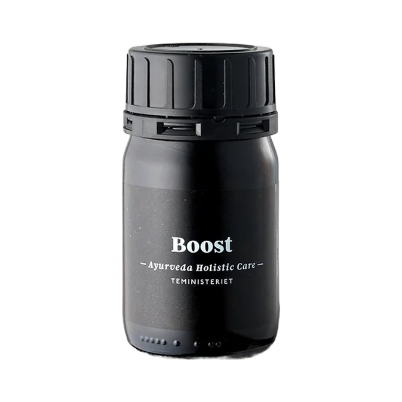 Glas med Ayurveda Boost Te. Sort tebeholder med teksten "Boost – Ayurveda Holistic Care" fra Teministeriet. Beholderen er cylindrisk med en sort skruelåg og minimalistisk design.