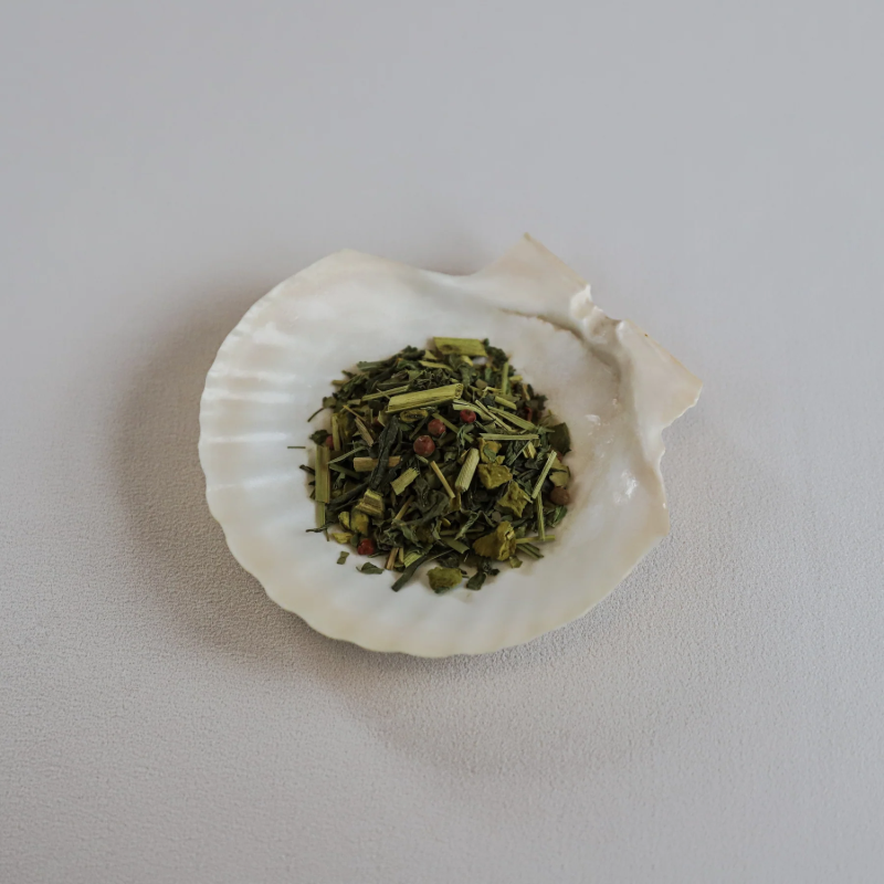 Løse teblade fra Teministeriet Ayurveda Boost te præsenteret i en hvid muslingeskal. Teen består af grønne yerba mate-blade, citrongræs, små røde peberkorn og tørrede citrusskaller. Muslingeskallen ligger på en lys overflade, og billedet har en naturlig og æstetisk præsentation, der fremhæver teens friske, let krydrede karakter.