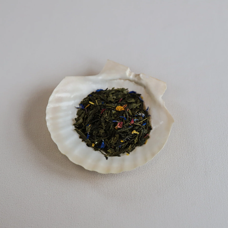 En nærbillede af den løse Green Sweet Bergamot-te fra Teministeriet, smukt anrettet i en lys muslingeskal. De lange, grønne teblade er blandet med farverige blomsterblade i blå, gule og røde nuancer, hvilket understreger teens friske og frugtige karakter.