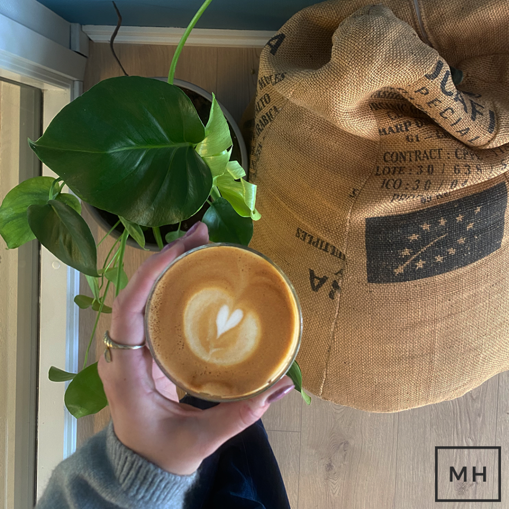 En person holder en kop kaffe i hånden. Under koppen ses en plante og en sækkepude lavet af gamle kaffeposer