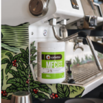 Beholder med Cafetto MFC Powder Eco rengøringsmiddel placeret foran en espressomaskine med dekorativt, planteinspireret design og dampstav i rustfrit stål.