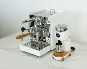 Hvid espressomaskine med træhåndtag og skinnende ståldetaljer fra Rocket står på en bordplade ved siden af en matchende hvid kaffekværn fra mærket Niche med træbase og metalbeholder.