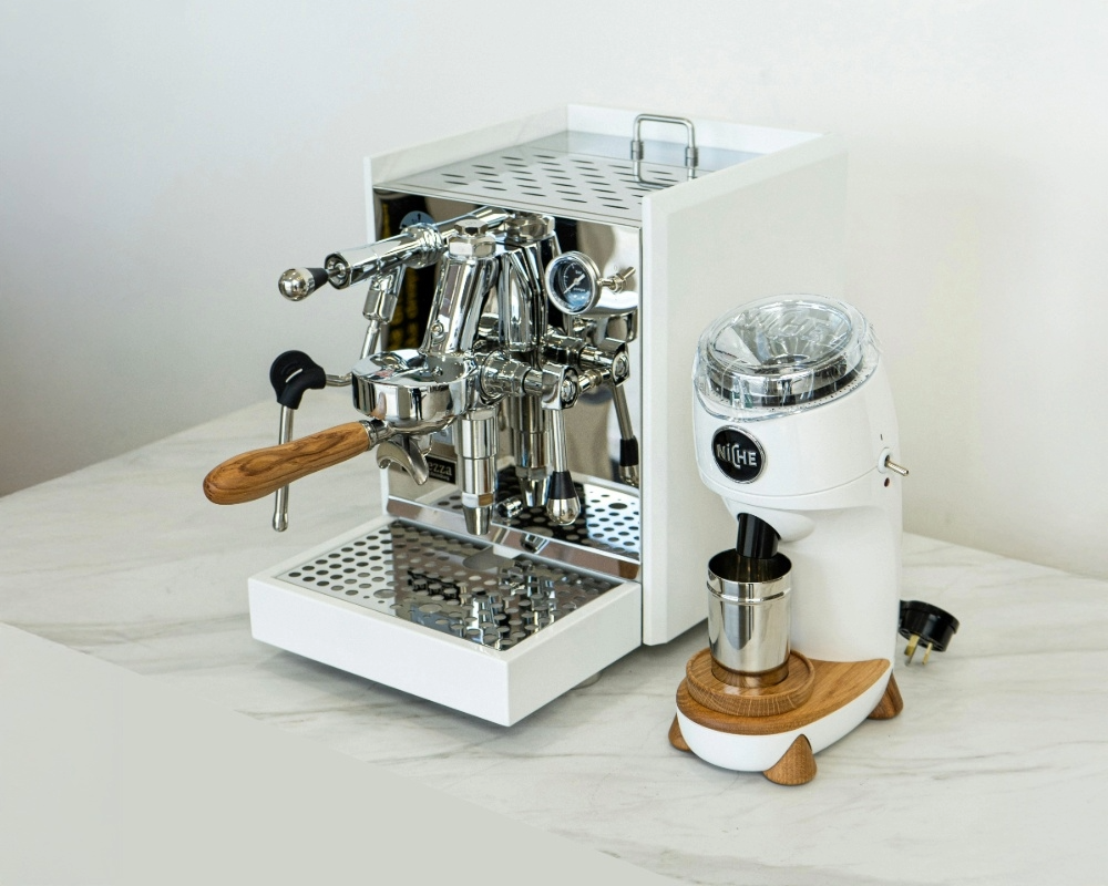 Hvid espressomaskine med træhåndtag og skinnende ståldetaljer fra Rocket står på en bordplade ved siden af en matchende hvid kaffekværn fra mærket Niche med træbase og metalbeholder.