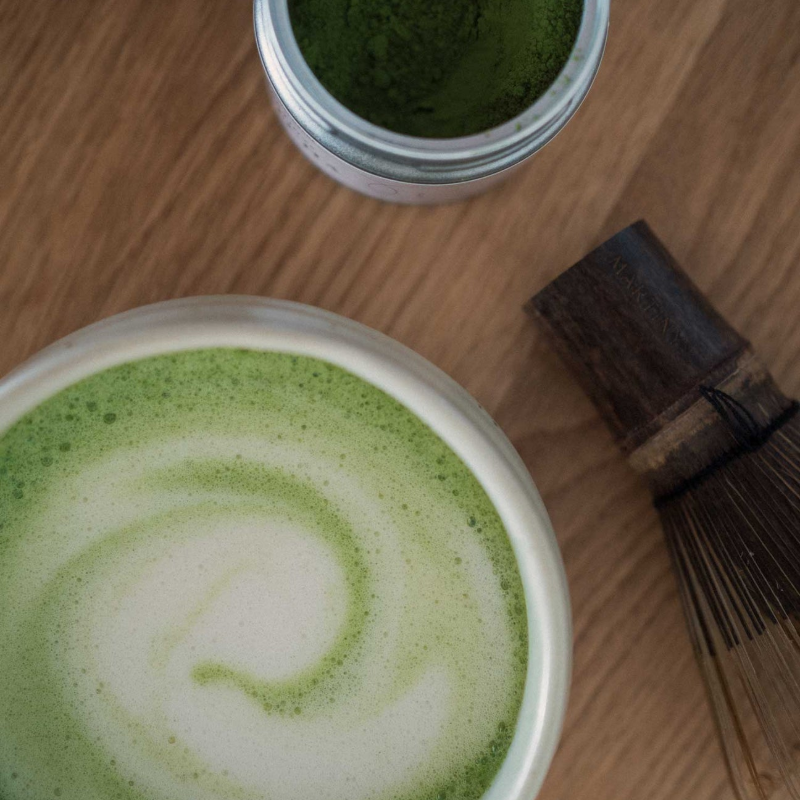 Topvinkel af en matcha latte med let hvid skum-s swirl i skålen; Martinys-dåse og bambuspiske ligger ved siden af på et træbord — varmt, naturligt stemningsfoto.