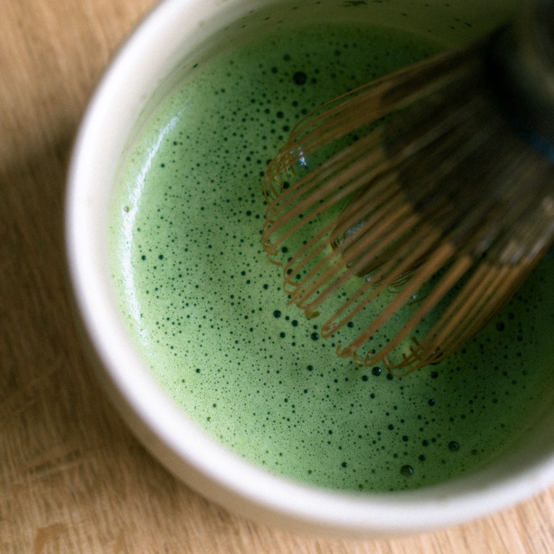 Tæt topbillede hvor en bambuspiske pisker grøn matcha i en skål; fokus på skummet under pisken og bevægelsen i den frodige, grønne overflade.