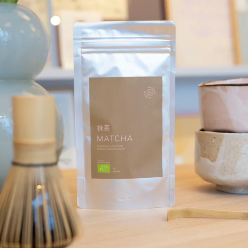Økologisk matcha te i pose placeret på bord ved siden af matcha-tilbehør og keramikkopper.