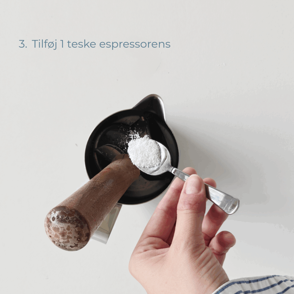 Hånd tilføjer en teske espressorens til mælkekanden med portafilter og filterkurv.