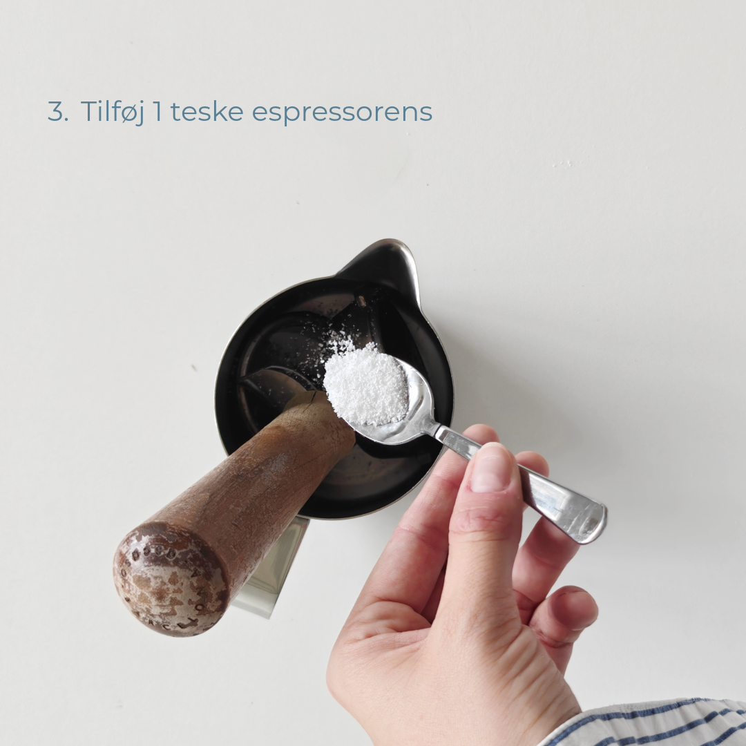 Hånd tilføjer en teske espressorens til mælkekanden med portafilter og filterkurv.