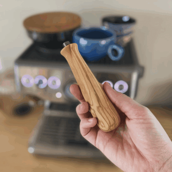 Udskifteligt håndtag i oliventræ til portafilter, vist i hånd foran Sage espressomaskine.