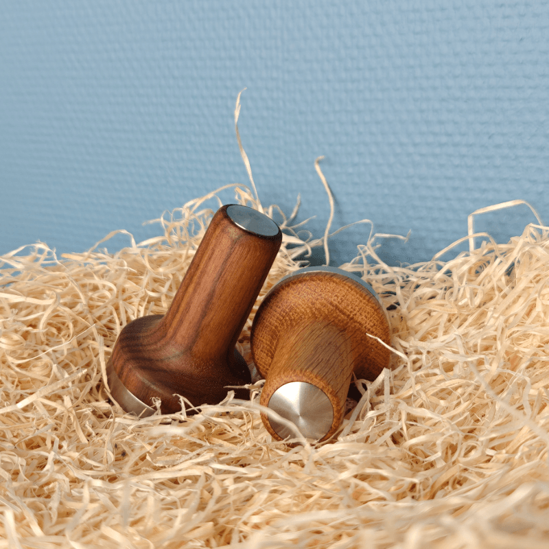Baark The Sage Tamper i valnød og eg – to håndlavede kaffetampere med stålbase placeret i træfyld mod blå baggrund.
