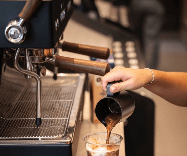 Til venstre ses en Ascaso espressomaskine. Til højre for den ses en hånd, som hælder espresso ned i en islatte.