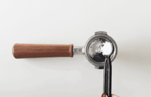 Portafilter med træhåndtag, hvor en ske med espressorens hældes i filterkurven – illustration af backflushing af espressomaskine.