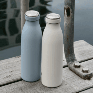 Miljøbillede af to AYA&IDA drikkeflasker stående på en træbro ved vandet — én i støvet blå og én i winter white — der viser flaskernes form og anvendelse udendørs.