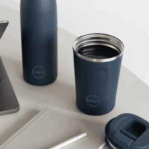 Stemningsbillede af AYA&IDA Cup2Go i mørkeblå stående på et bord ved en arbejdsstation, låg taget af og et matchende termoflaske i baggrunden; viser krusets indre og praktiske størrelse.