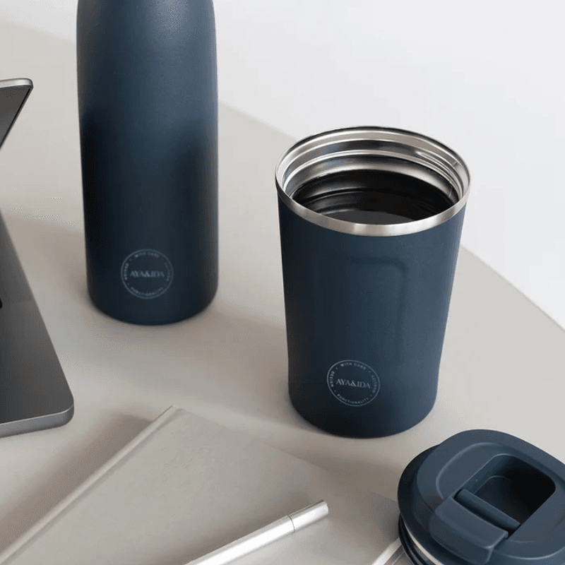 Stemningsbillede af AYA&IDA Cup2Go i mørkeblå stående på et bord ved en arbejdsstation, låg taget af og et matchende termoflaske i baggrunden; viser krusets indre og praktiske størrelse.