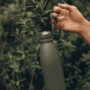 AYA&IDA drikkeflaske 1000 ml i tropical green hængende i en bærerem holdt af en hånd foran grønne planter — viser flaskens bærbarhed og udendørsbrug.