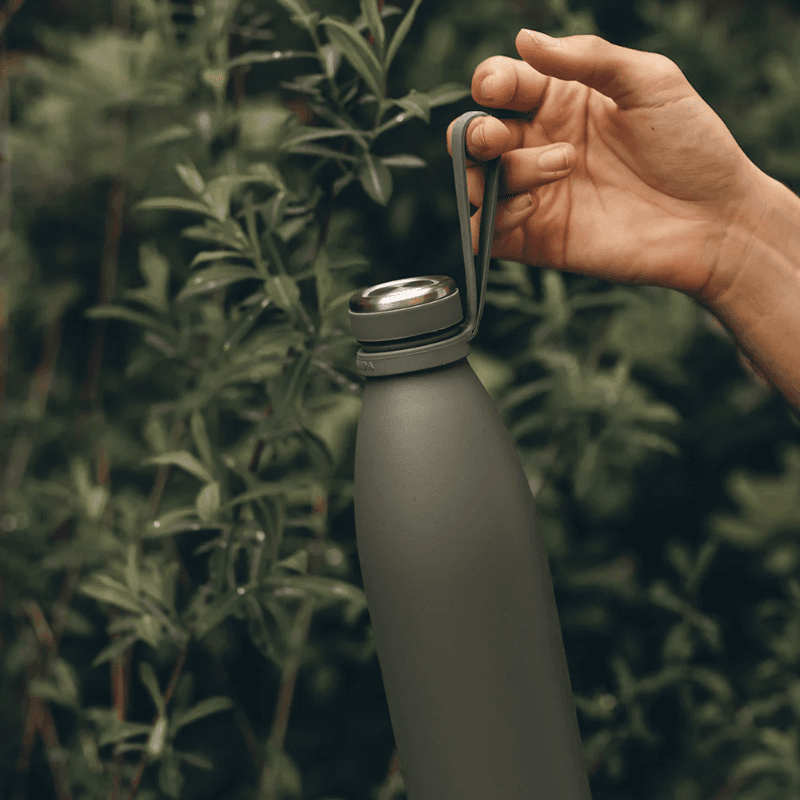 AYA&IDA drikkeflaske 1000 ml i tropical green hængende i en bærerem holdt af en hånd foran grønne planter — viser flaskens bærbarhed og udendørsbrug.