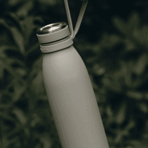 Miljøfoto af AYA&IDA bærerem/hank til drikkeflaske vist på en flaske og holdt i remmen foran grønne planter — viser praktisk brug og bæreevne udendørs.