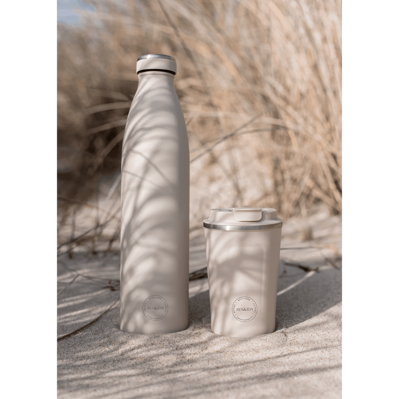 AYA&IDA drikkeflaske 1000 ml og Cup2Go termokrus i creme/beige med mat finish og sølvkant placeret i sandet foran tørre strandgræsser; bløde skygger fra sivene skaber en rolig, naturlig stemning.