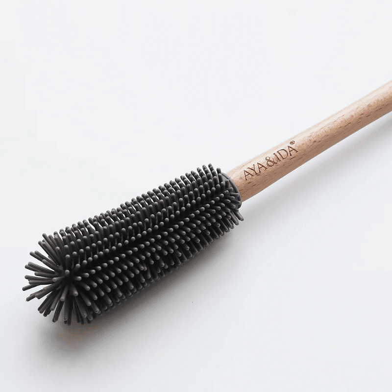 Nærbillede af AYA&IDA flaskebørste der ligger på hvid overflade; tydeligt træskaft med indgraveret logo og mørkegråt silikonehoved med fleksible børster.