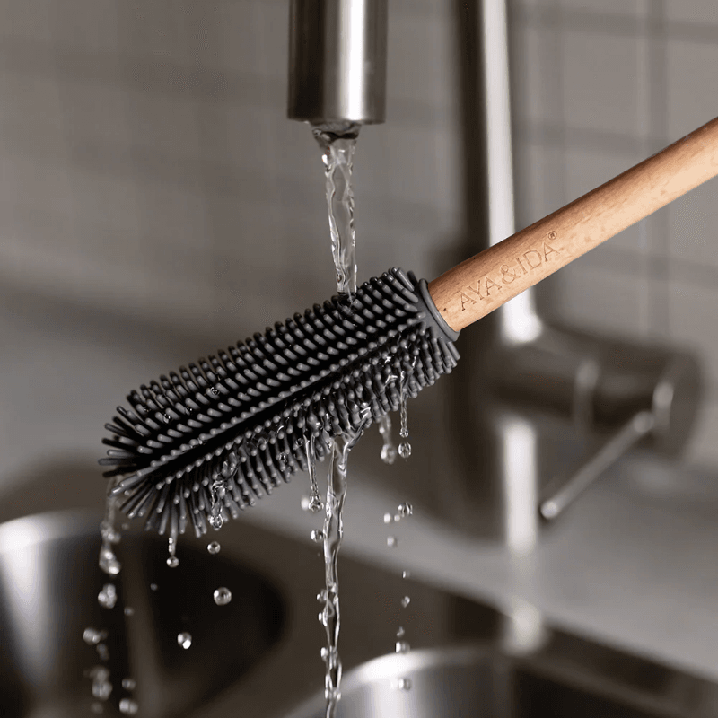 AYA&IDA flaskebørste holdt under rindende vand i køkkenvasken; vanddryp og silikonebørster vist for at illustrere nem rengøring og aftørring.