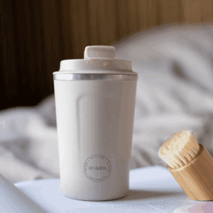 AYA&IDA Cup2Go - 500 ml termokrus i creme/beige med mat overflade og sølvkant, lukket kliklåg med en lille dråbe, placeret på en bog ved siden af en bambusbørste foran en blød, udtonet baggrund (senge-/hjemmestemning).