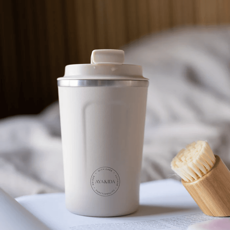 AYA&IDA Cup2Go - 500 ml termokrus i creme/beige med mat overflade og sølvkant, lukket kliklåg med en lille dråbe, placeret på en bog ved siden af en bambusbørste foran en blød, udtonet baggrund (senge-/hjemmestemning).