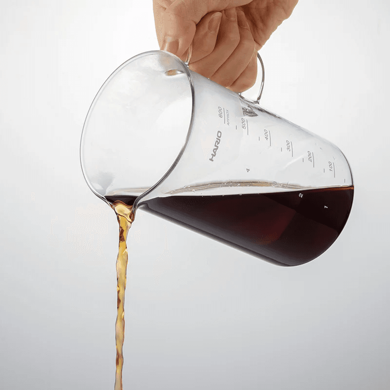 Hario Beaker Server 600 ml i klart glas set i skrå vinkel mens friskbrygget sort kaffe hældes fra tuden; synlig måleskala og hank.