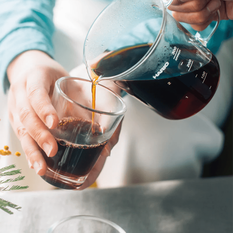 Stemningsbillede af Hario Beaker Server med en Hario V60 keramisk dripper på toppen og friskbrygget kaffe i beakeren; bløde baggrundsdetaljer med mælkekande i baggrunden.