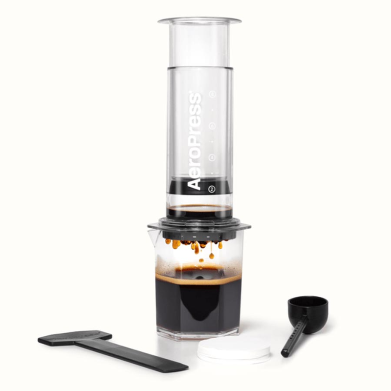 AeroPress XL Clear i gennemsigtig plast monteret på måleglas, ved siden af følger med skovl, rørepind og papirfiltre; et færdigbrygget krus står klar på bordet.