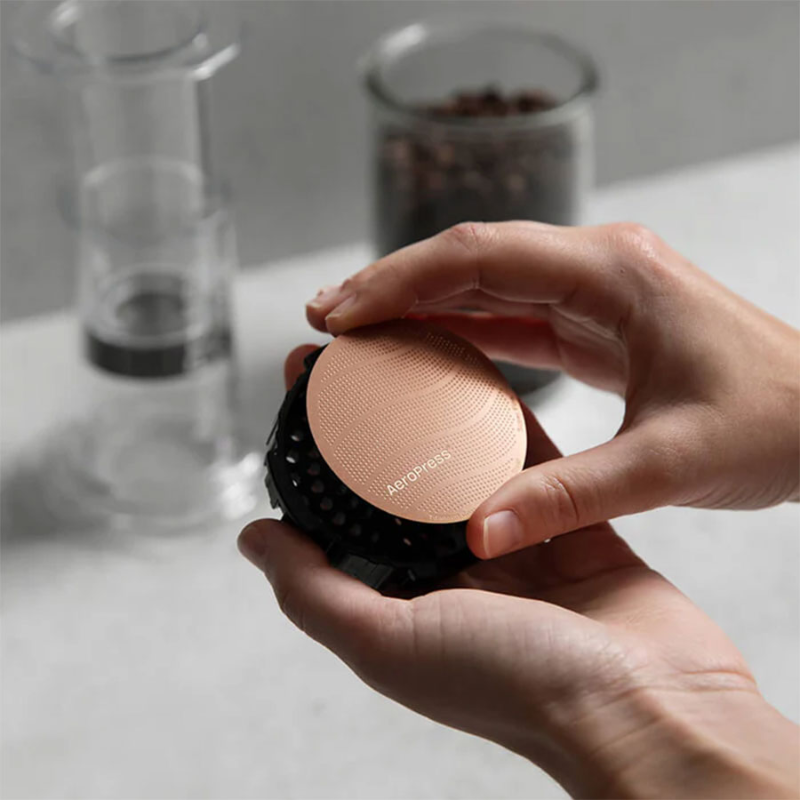 AeroPress Gold Tone Metalfilter — hænder monterer det kobberfarvede metalfilter i AeroPress’ plastindsats, viser hvordan filteret sættes på plads.