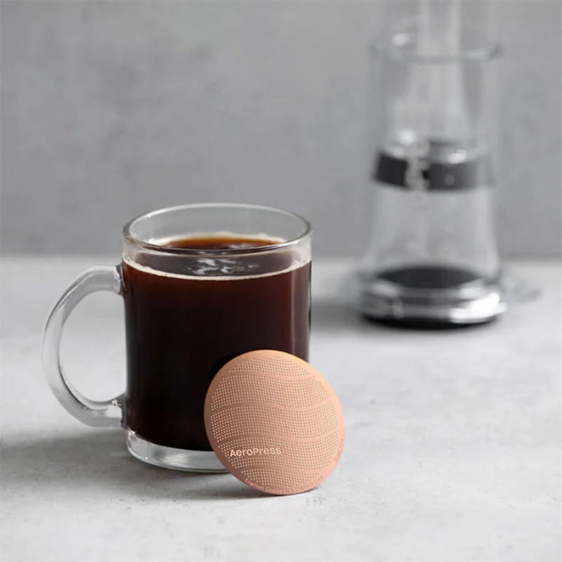 AeroPress Gold Tone Metalfilter — kobberfarvet metalfilter liggende foran en krus med sort kaffe og en AeroPress i baggrunden, stemningsbillede fra køkkenet.